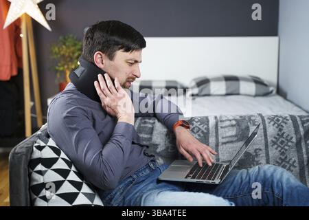 Man employee after accident working at home behind laptop and feels pain in neck, uses black neck collar support. Male with neck injury freelance prog Stock Photo