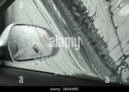 View from inside of car to side mirror and window being washed in carwash, brush leaving strokes in soap foam and shampoo Stock Photo