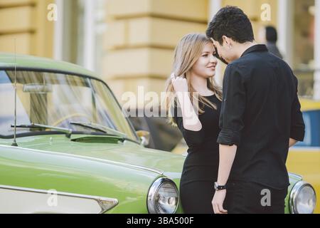 Young multiracial couple, male and female lovers heterosexual people students. Beautiful models posing standing near a retro car in the city. Dressed Stock Photo