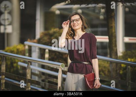 Caucasian adult lady woman in glasses posing near office building outside. Business lady in skirt holds hand glasses for vision bag in hand. Professio Stock Photo