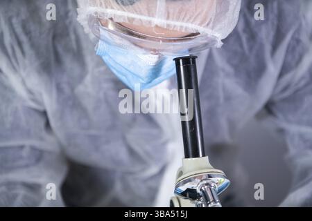 Medical professional in white protective suit looking through microscope in chemical laboratory. Search vaccine coronavirus. Covid 19 drug inventions. Stock Photo