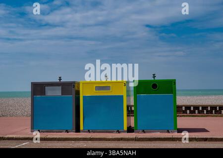 Three trash cans are lined up on the sidewalk, one of which is green. The other two are blue and yellow. The scene is set against a backdrop of a blue Stock Photo