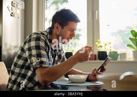 Man in plaid shirt eating oatmeal porridge from white bowl while using smartphone. Male looking at phone during breakfast. Concept of social media add Stock Photo