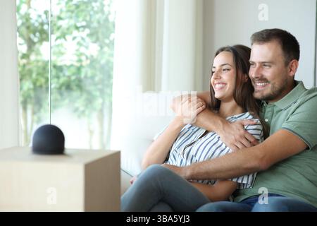 Happy couple with smart speaker enjoying sitting on a couch at home Stock Photo