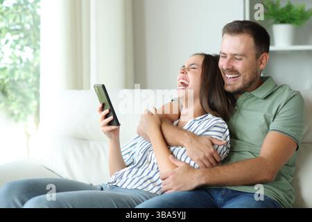 Happy couple laughing loud checking cell phone content sitting on a couch at home Stock Photo