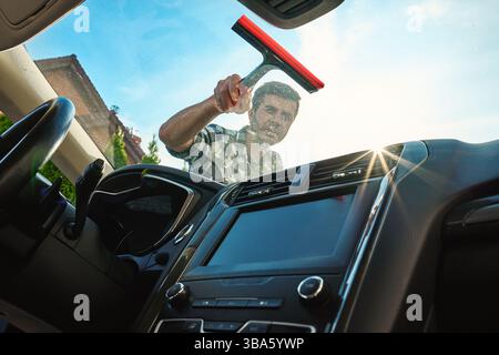 Male washing windshield in sunlight, view from from car inside. Man cleaning car windshield from outside using squeegee. Concept of car care, cleaning Stock Photo