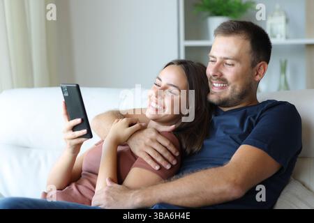 Happy couple using smart phone hugging sitting on a sofa at home Stock Photo