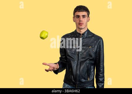 Young man wearing black leather jacket juggling green apple on yellow background Stock Photo