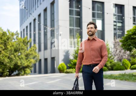 A smiling businessman walks confidently towards the viewer, carrying a briefcase. The scene is set outdoors near a modern office building. Stock Photo