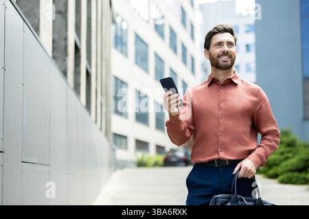 A well-dressed man walks outside, holding a phone in one hand and a briefcase in the other, smiling. Stock Photo