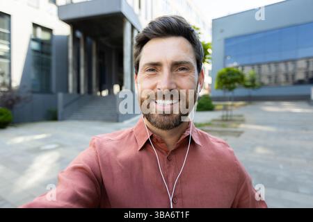 A man smiles during a video call, holding his phone. He wears earphones while standing in an urban outdoor environment Stock Photo