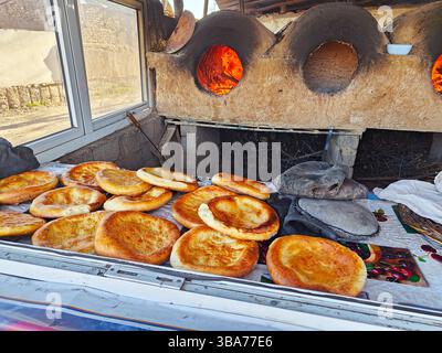 Breads are cooling on a table in front of a traditional stone oven. The setting is rural, with glowing embers visible and sun illuminating the scene. Stock Photo