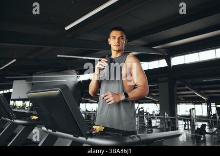 Active young guy in grey tshirt drinking water from bottle, standing on treadmill. Stock Photo