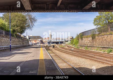 A railway station viewed from under a metal bridge. Rail tracks lead to the station building with platforms and a clock tower. A sky with cloud is abo Stock Photo