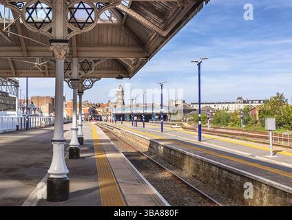A railway station viewed from under a historic canopy with columns. Rail tracks lead to the station building with platforms and a clock tower. A sky w Stock Photo