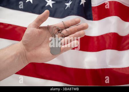 Mans hand holds military soldiers tag against backdrop of American flag. National pride and military tribute with focus on tag. Memorial Day. Stock Photo