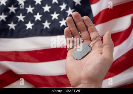 Mans hand holds military soldiers tag against backdrop of American flag. National pride and military tribute with focus on tag. Memorial Day. Stock Photo