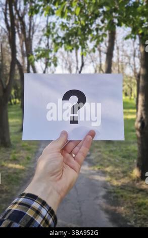 Male hand holding a white sheet of paper with a question mark on a path in the woods. Curiosity and uncertainty symbol. Lost person, confusion concept Stock Photo