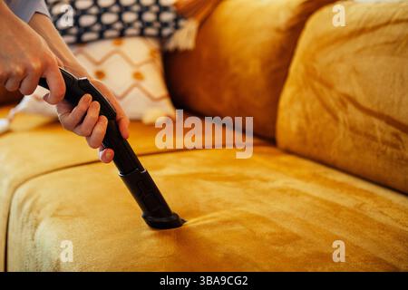 Close up of a female hands with a suction brush vacuuming the upholstery of a sofa. A woman cleans a couch using a vacuum cleaner and a nozzle for uph Stock Photo