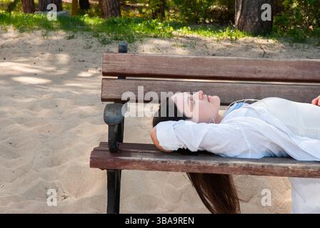 Young caucasian woman relaxing on wooden park bench at sandy beachside, enjoying peaceful summer afternoon under shade of trees. Digital nomad lifesty Stock Photo