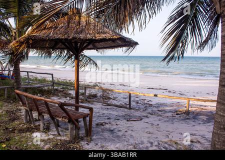 A rustic wooden bench under a thatched umbrella on the beach at the Inhassoro in Mozambique. Stock Photo