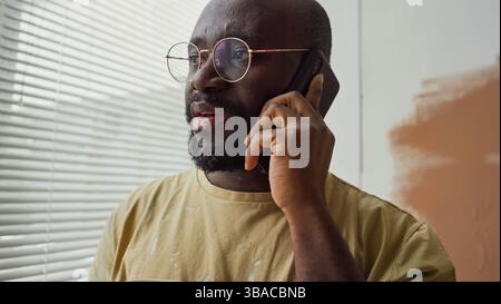 Man standing near window blinds, holding phone and looking concerned, wearing glasses and casual shirt, with partially painted wall in background Stock Photo