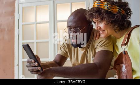 Couple smiling and looking at smartphone inside home with natural lighting coming through window. Man in casual t-shirt and woman in patterned headband both seem engaged with screen Stock Photo