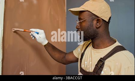 African American painter concentrating on painting wall using brush while wearing cap and overalls. Worker seen in profile focusing on task with paintbrush in hand Stock Photo