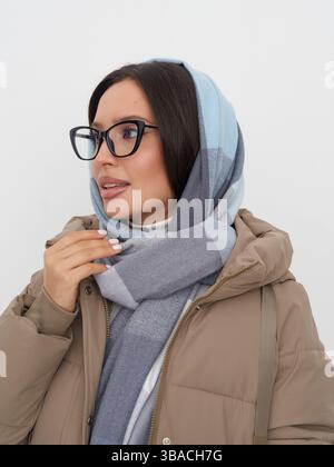 Woman with glasses wears a light blue and grey scarf and a beige jacket. Her dark hair is partially covered by the scarf as she gazes to the side with Stock Photo