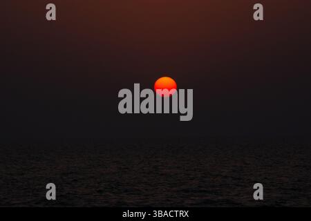 A vibrant sunset over the ocean, with a large, glowing orange sun setting against a dark, hazy sky. The water reflects the warm colors of the sun, cre Stock Photo