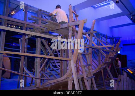 A wooden shipbuilding exhibit featuring a person working on the structure. The scene is illuminated with blue lighting, highlighting the intricate woo Stock Photo