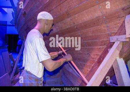 A craftsman working on a wooden boat, using tools to shape the wood. The scene is illuminated with blue lighting, highlighting the craftsmanship invol Stock Photo