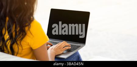 Unrecognizable black lady typing on laptop with blank screen on comfy couch at home, mockup for design Stock Photo