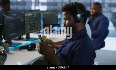 Admin in server room using mobile phone, relaxing after ensuring minimal downtime. Data center IT expert texting on smartphone, taking break from monitoring performance, camera A Stock Photo