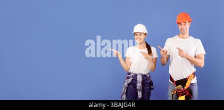 Smiling handyworkers couple wearing casual clothes and protective hard hats pointing aside Stock Photo