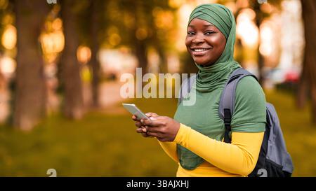 Outdoor Portrait Of Happy Black Muslim Female Student With Smartphone And Backpack Stock Photo