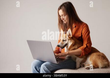 Young woman working remotely with her Corgi dog sitting on lap, using laptop at cozy home office. Stock Photo