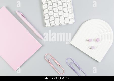 Top view of white keyboard, pink notebook, pink pen on grey background. Paper clips, pins, modern office, school. White arch tray, flat lay. Stock Photo