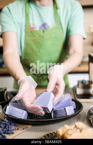 Woman in green apron presenting freshly crafted lavender soap bars on a plate with gloved hands. Handmade wellness, chemical-free skincare, female Stock Photo