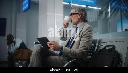 Airport Terminal: Successful Entrepreneur Using Digital Tablet and Drinking Coffee, Doing e-Business. Businessman Remote Work Online Waiting for Flight in Departure Lounge. Slow Motion, Medium Shot. Stock Photo