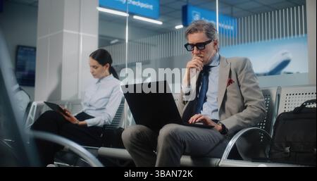 VIP Departure Lounge in International Airport Terminal: Mature Businessman Using Laptop, Sending e-Commerce Emails. Diverse Businesspeople Working with Digital Devices Waiting for Flight. Dolly Shot. Stock Photo