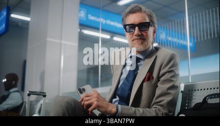 Airport Terminal: Mature Businessman Using Smartphone and Drinking Coffee, Looking at Camera. Entrepreneur with Luggage Waiting for Flight in Departure Lounge, Going on Business Trip. Dolly Shot. Stock Photo
