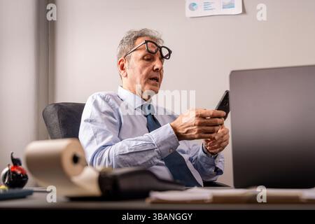 Mature executive in shirt and tie reacts to bad news on his phone while sitting in front of a laptop; concept of bill shock, banking problem, cost ove Stock Photo