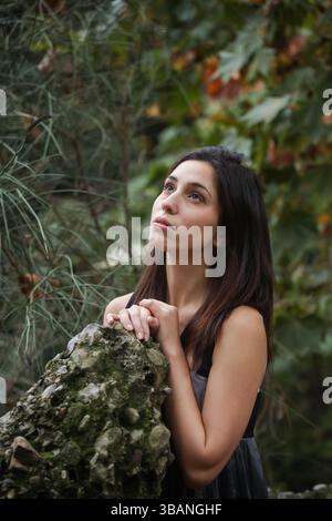 Dreamy young woman leaning on rock, surrounded by vibrant green and autumn leaves. Stock Photo