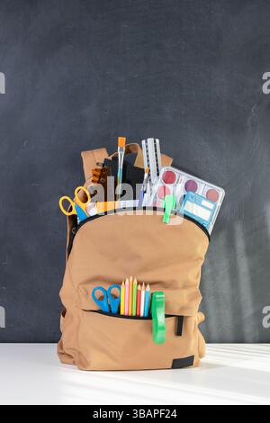 School backpack overflowing with Stationery Supplies on blackboard with copy space, ready for back to school season Stock Photo