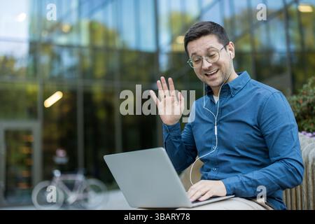 A man is sitting outside, using a laptop while waving to someone. He is smiling and appears to be having a video call. Stock Photo