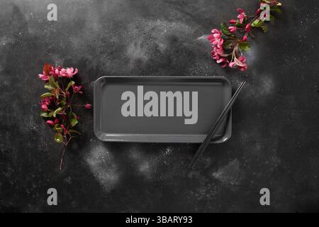 Minimalist scene of empty rectangular plate for asian food with black chopsticks on dark textured surface with spring pink flowers. Asian style. View Stock Photo