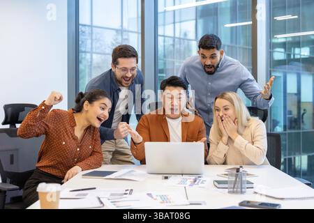 A diverse team celebrates a successful project, looking excitedly at a laptop screen. Teamwork and achievement are evident. Stock Photo