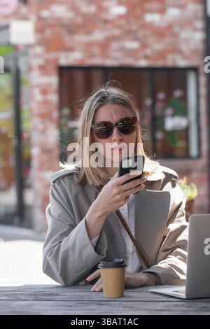 Young woman working using phone and laptop sitting on cafe veranda. Remote work, businesswoman Stock Photo