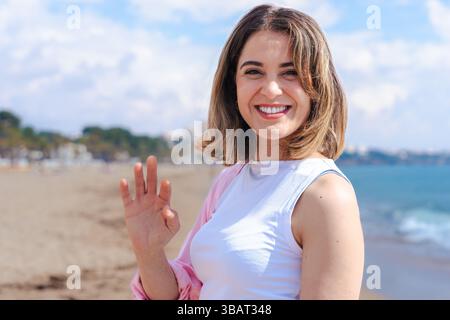 Smiling Woman Waving Hand at Camera While Standing on Sunny Beach Near Ocean Stock Photo
